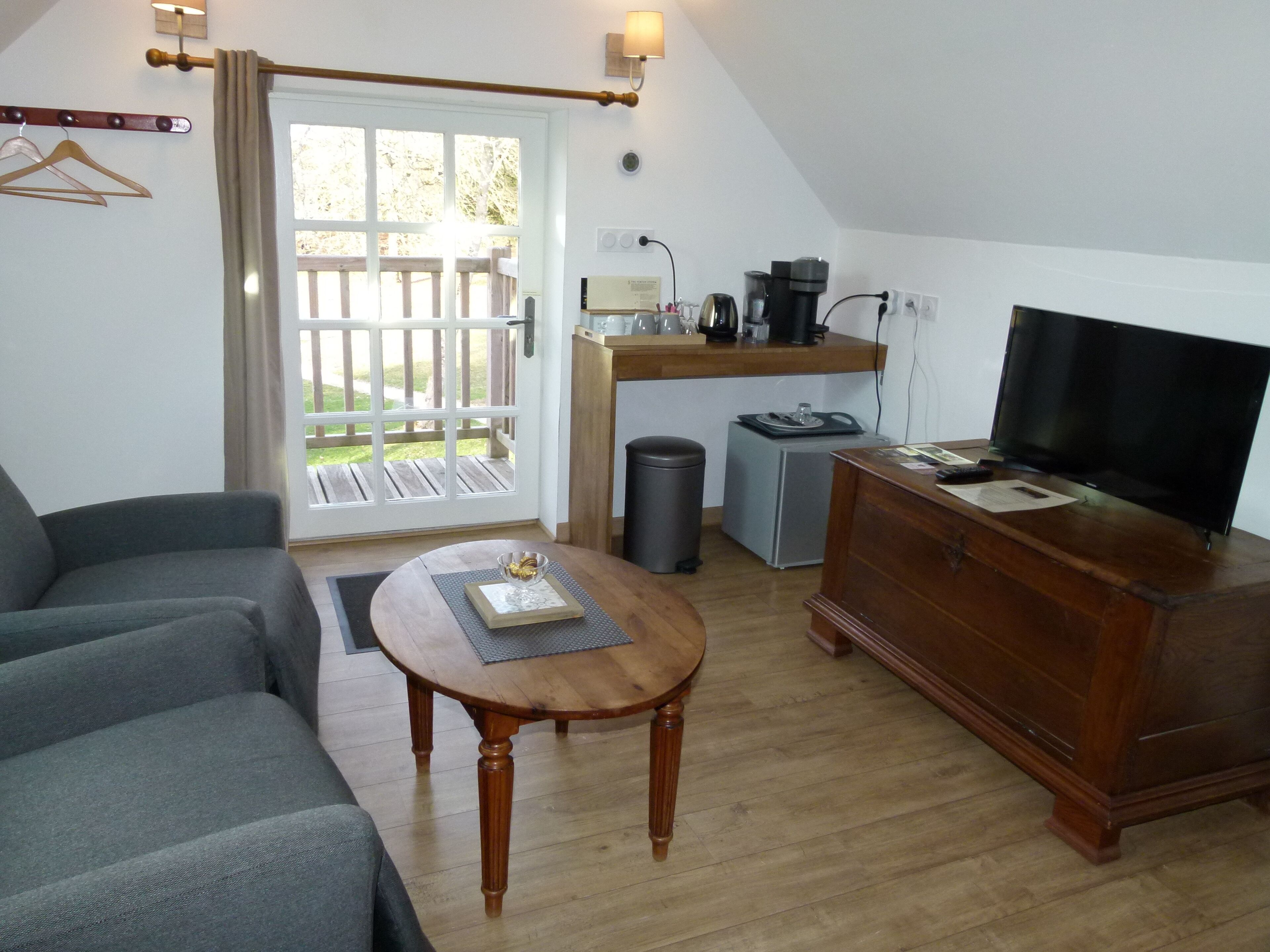 A cozy living room with a sofa, a coffee table, a TV, a small kitchen area with a coffee maker, and a trash bin.