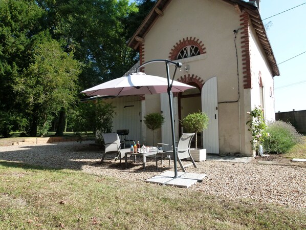 An outdoor patio with a table and chairs under a large umbrella, surrounded by gravel and grass, with a building in the background. - Le Grand Saint-Marc (Vendome)