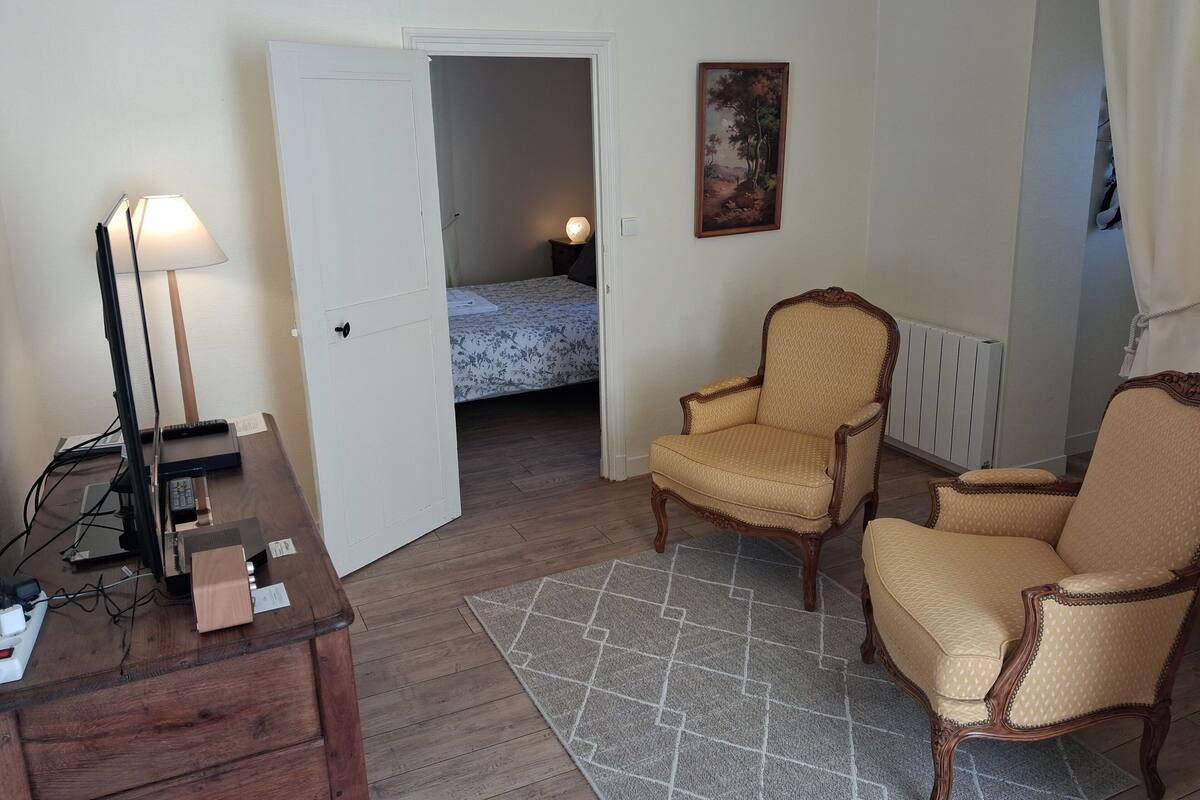 Ein Zimmer mit Schreibtisch, zwei Sesseln, einem Bett, einem Gemälde und einer Heizung.