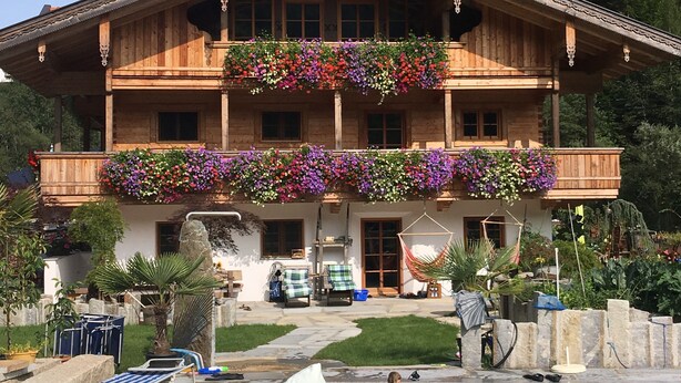 A wooden chalet with a balcony adorned with colorful flowers, a swimming pool, and a stone pathway leading to the entrance. - Haus Anni im Paradies (Kiefersfelden)