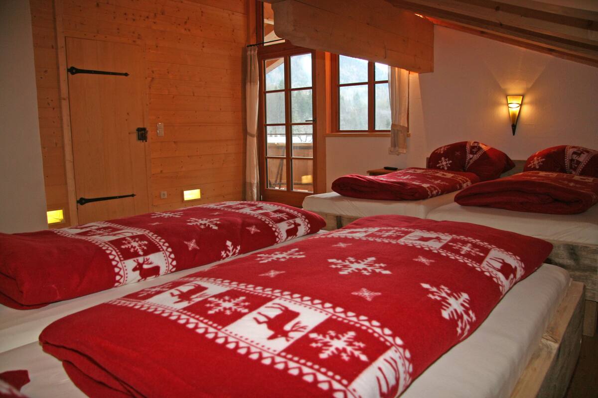 A room with wooden walls, two beds with red bedding, a window with curtains, and a wall-mounted light fixture.
