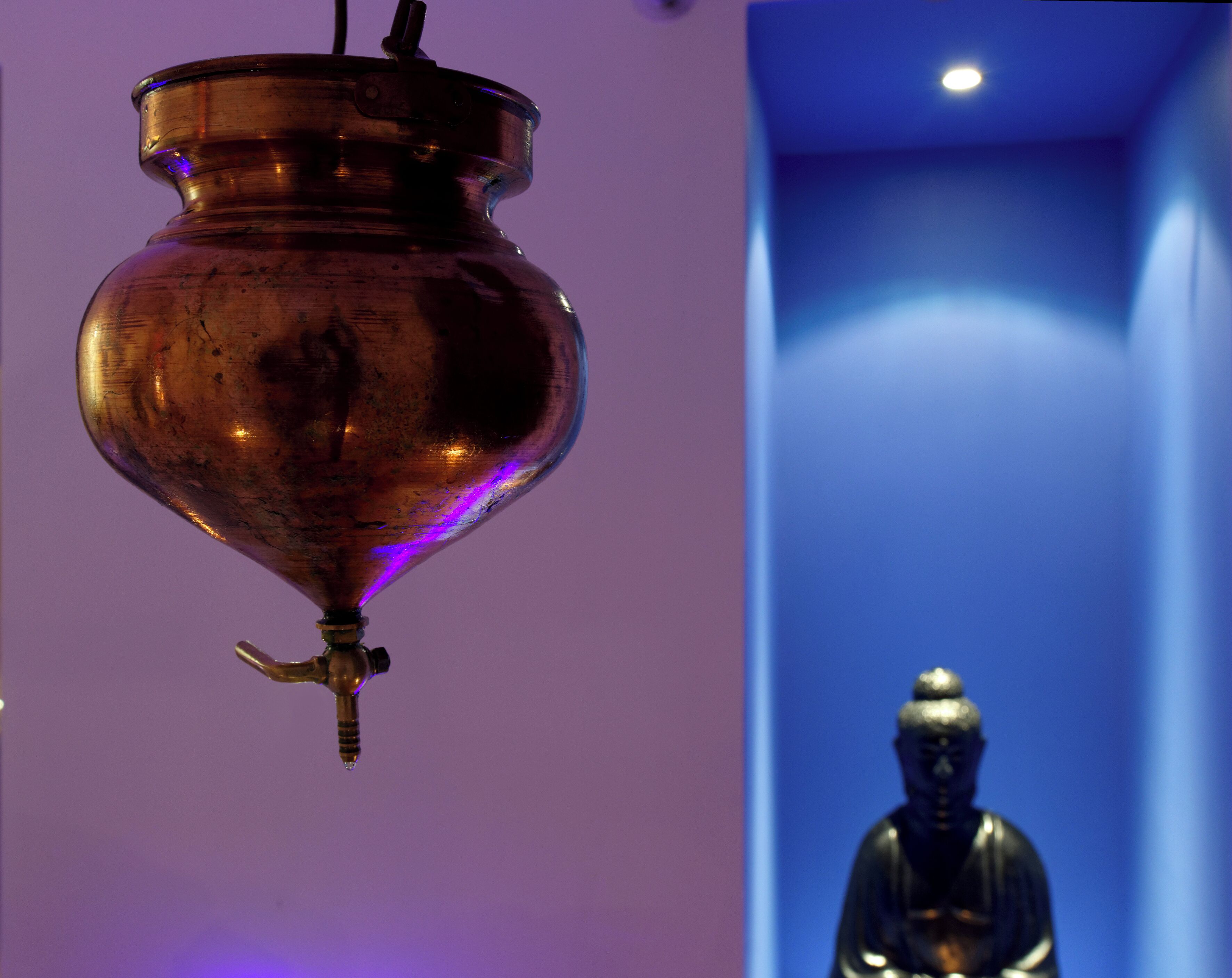 A hanging copper vessel with a spout, a statue of a seated figure, and a blue-lit corridor.