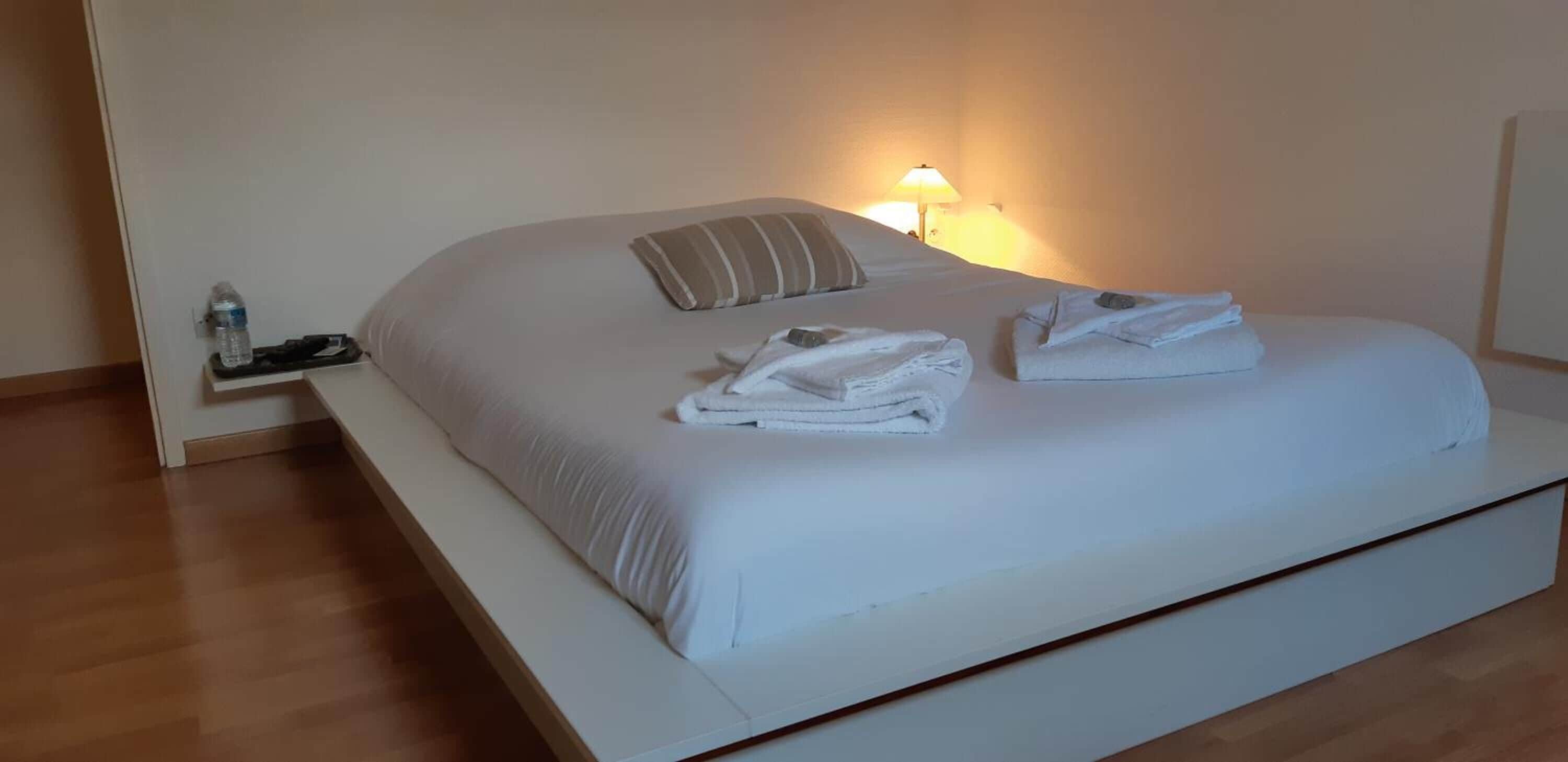 A neatly made bed with white linens and a striped pillow, a bedside table with a water bottle and a lamp, and a wooden floor.