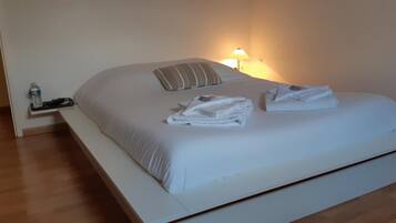 A neatly made bed with white linens and a striped pillow, a bedside table with a water bottle and a lamp, and a wooden floor.