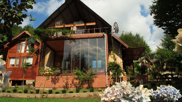A wooden cabin with a large glass window, surrounded by a garden with flowers and a clear sky. - Karaca Dag Evleri (Basiskele)