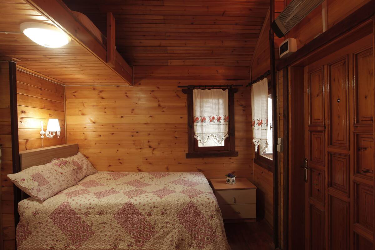 A wooden cabin bedroom with a bed, bedside lamp, nightstand, and a window with lace curtains.