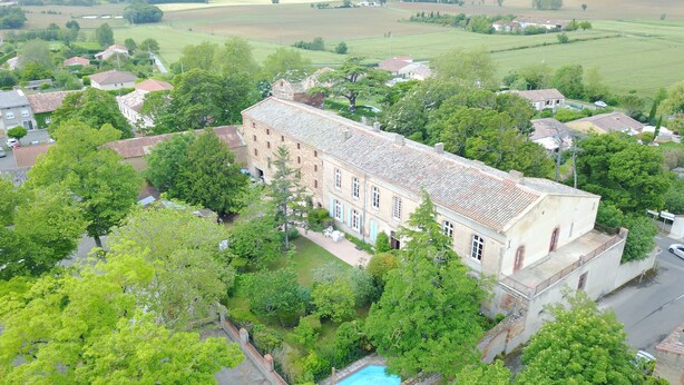 Un edificio grande e histórico con tejado de tejas, rodeado de abundante vegetación y una piscina.