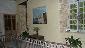 Un interior espacioso con techo alto, un balcón con barandilla de hierro forjado, un cuadro de un paisaje urbano en la pared y una ventana con vista a zonas verdes.