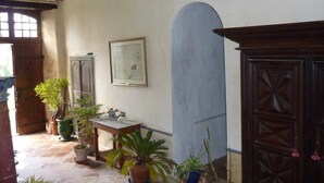 Una habitación con suelo de baldosas, una puerta de madera, una mesa con una planta y un cuadro en la pared.