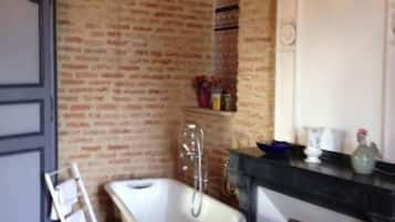 Un baño con una bañera de patas de león, una silla de madera, un perchero para toallas y una pared de ladrillo.
