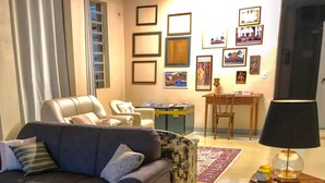 Un salon avec un canapé, des fauteuils, une table basse et un tapis. Des tableaux encadrés ornent le mur et une valise est posée sur le sol.