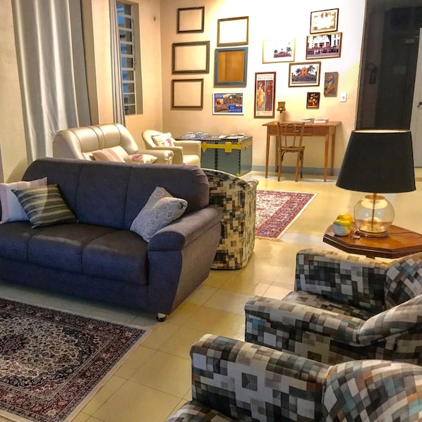Un salon avec un canapé, des fauteuils, une table basse et un tapis. Des tableaux encadrés ornent le mur et une valise est posée sur le sol.