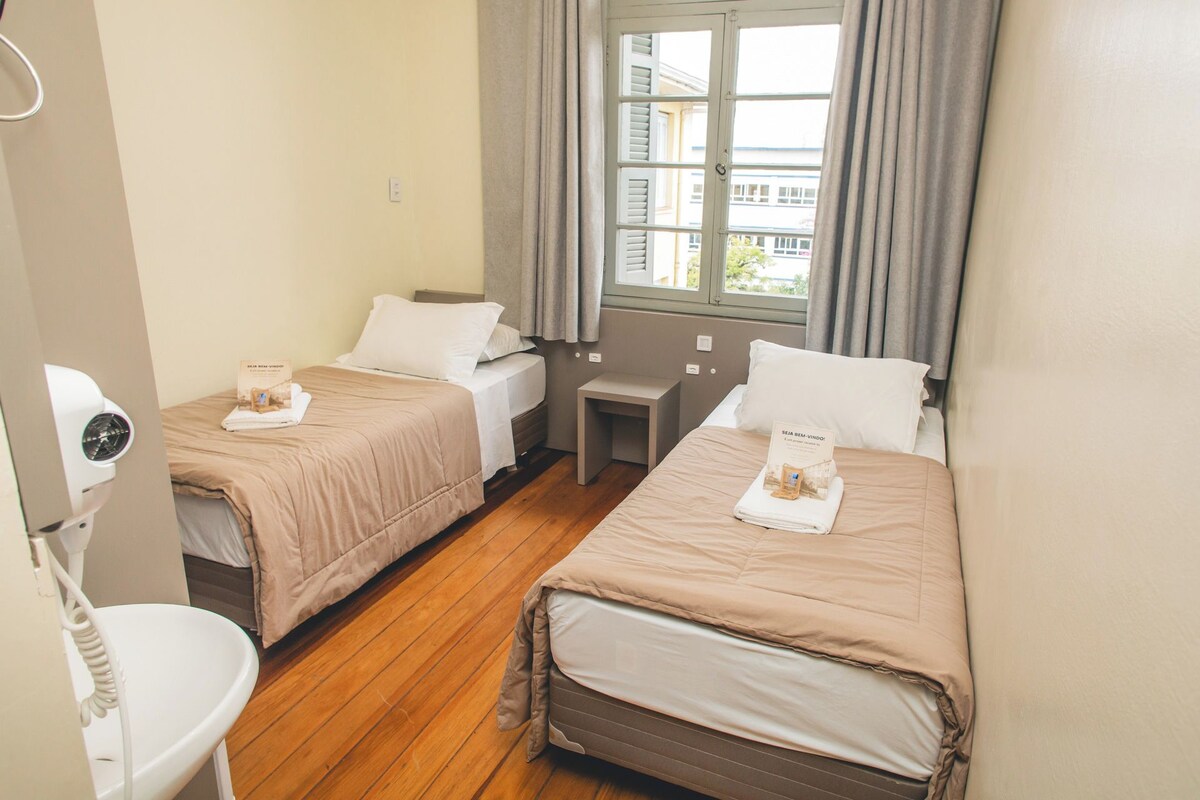 Une chambre d’hôtel avec deux lits, une petite table de chevet, une fenêtre avec des rideaux et une salle de bain avec des toilettes et un lavabo.