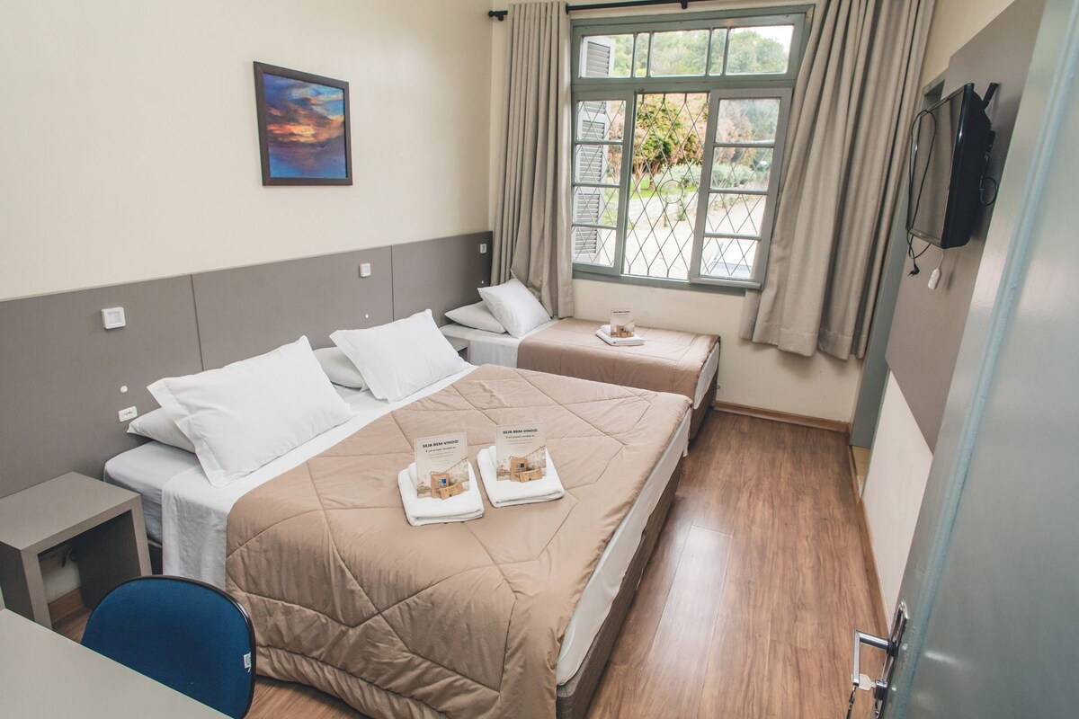 Une chambre d’hôtel avec deux lits, un bureau, une chaise, une fenêtre avec des rideaux et un tableau au mur.