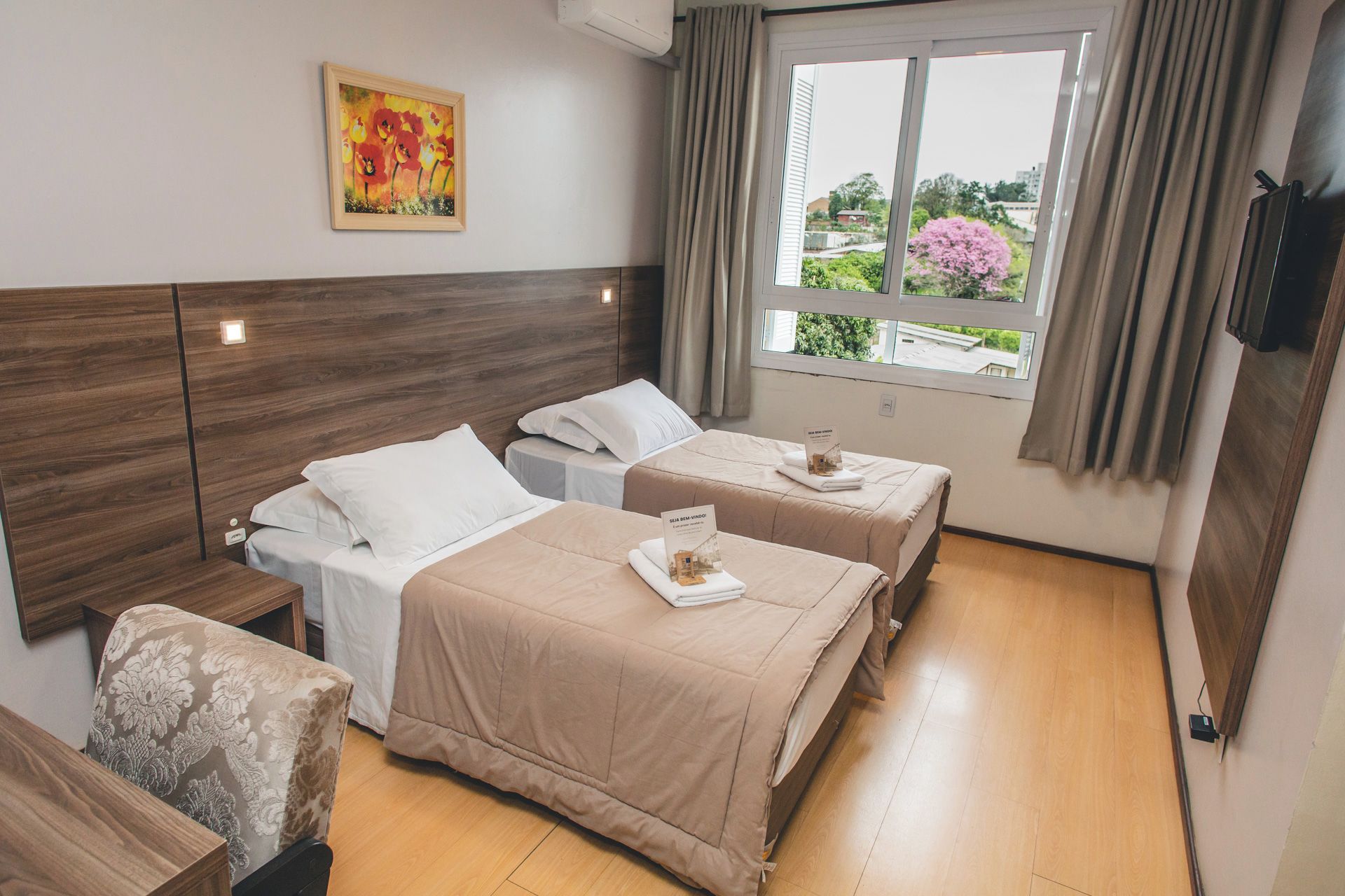 A hotel room with two beds, a wooden headboard, a painting, and a window with a view of greenery.