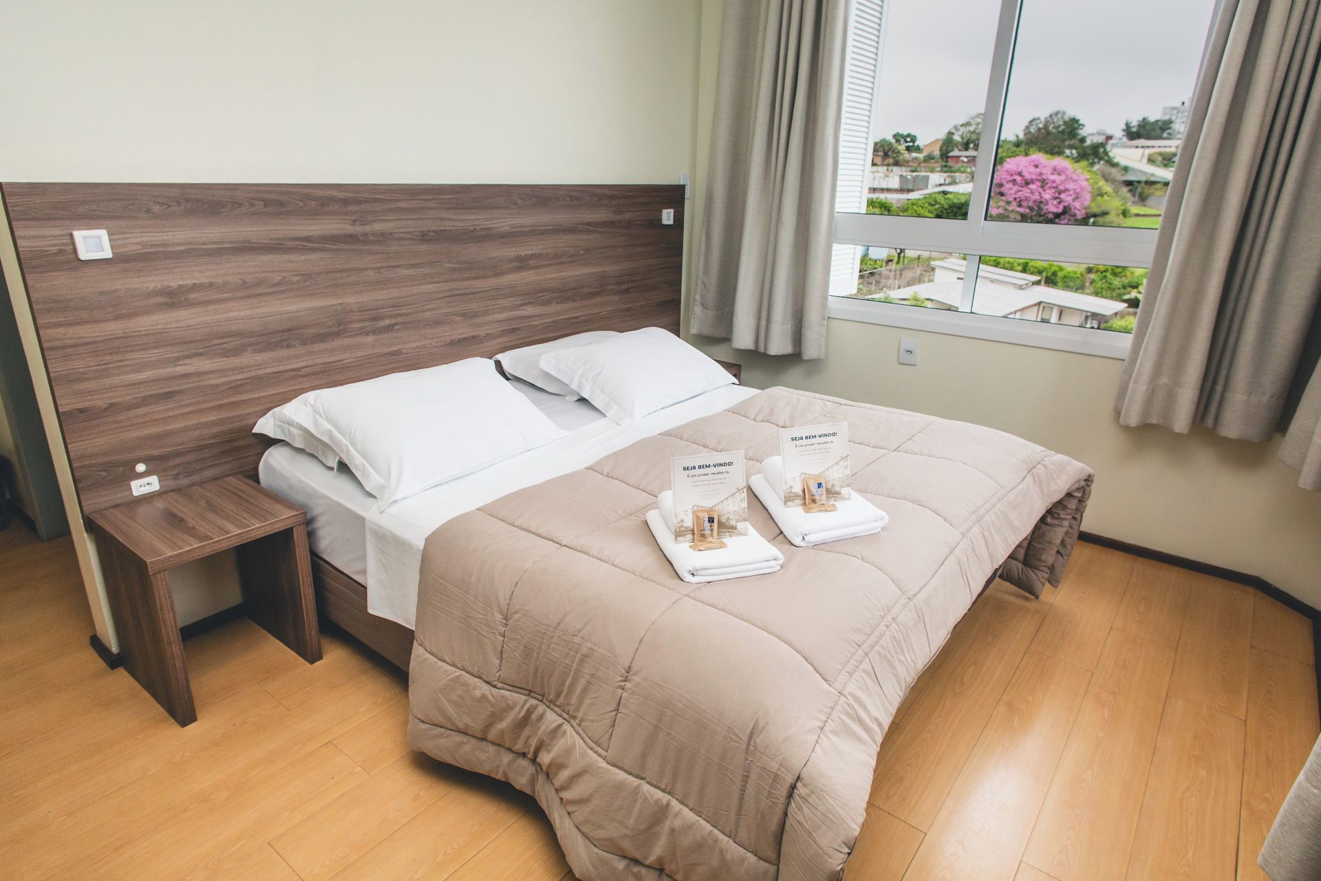 Cama bem arrumada com edredom bege e travesseiros brancos, cabeceira de madeira, mesa de cabeceira com tomada e janela com vista para vegetação e edifícios.