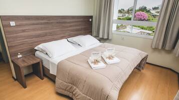 Cama bem arrumada com edredom bege e travesseiros brancos, cabeceira de madeira, mesa de cabeceira com tomada e janela com vista para vegetação e edifícios.