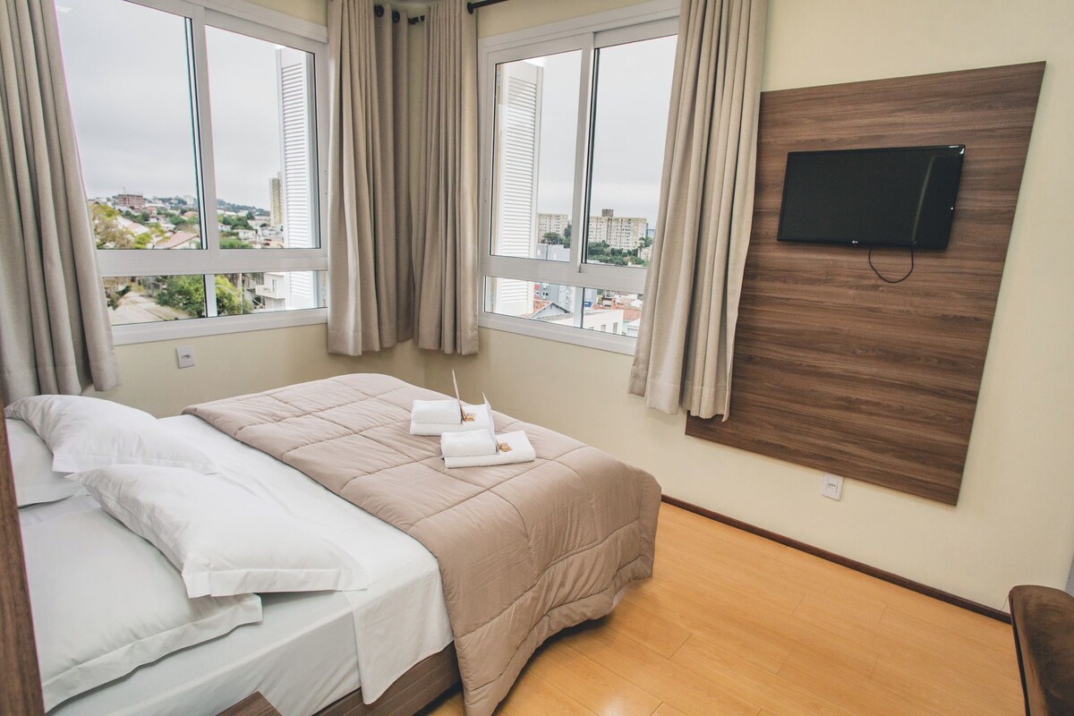 Une chambre d’hôtel avec un lit, un téléviseur fixé au mur et une vue sur la ville grâce à de grandes fenêtres.