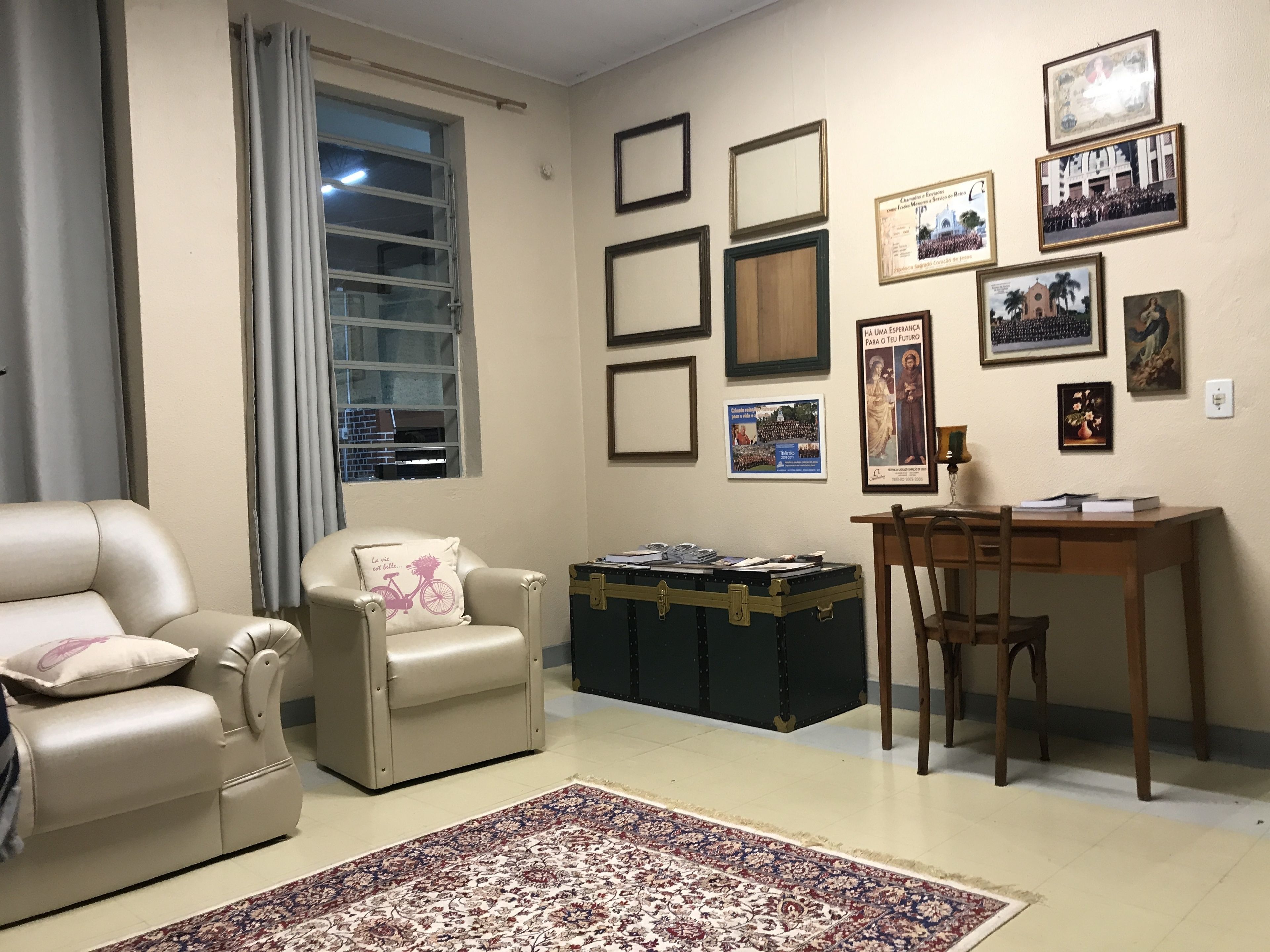 Uma sala de estar com sofá, uma poltrona, escrivaninha e uma janela grande.