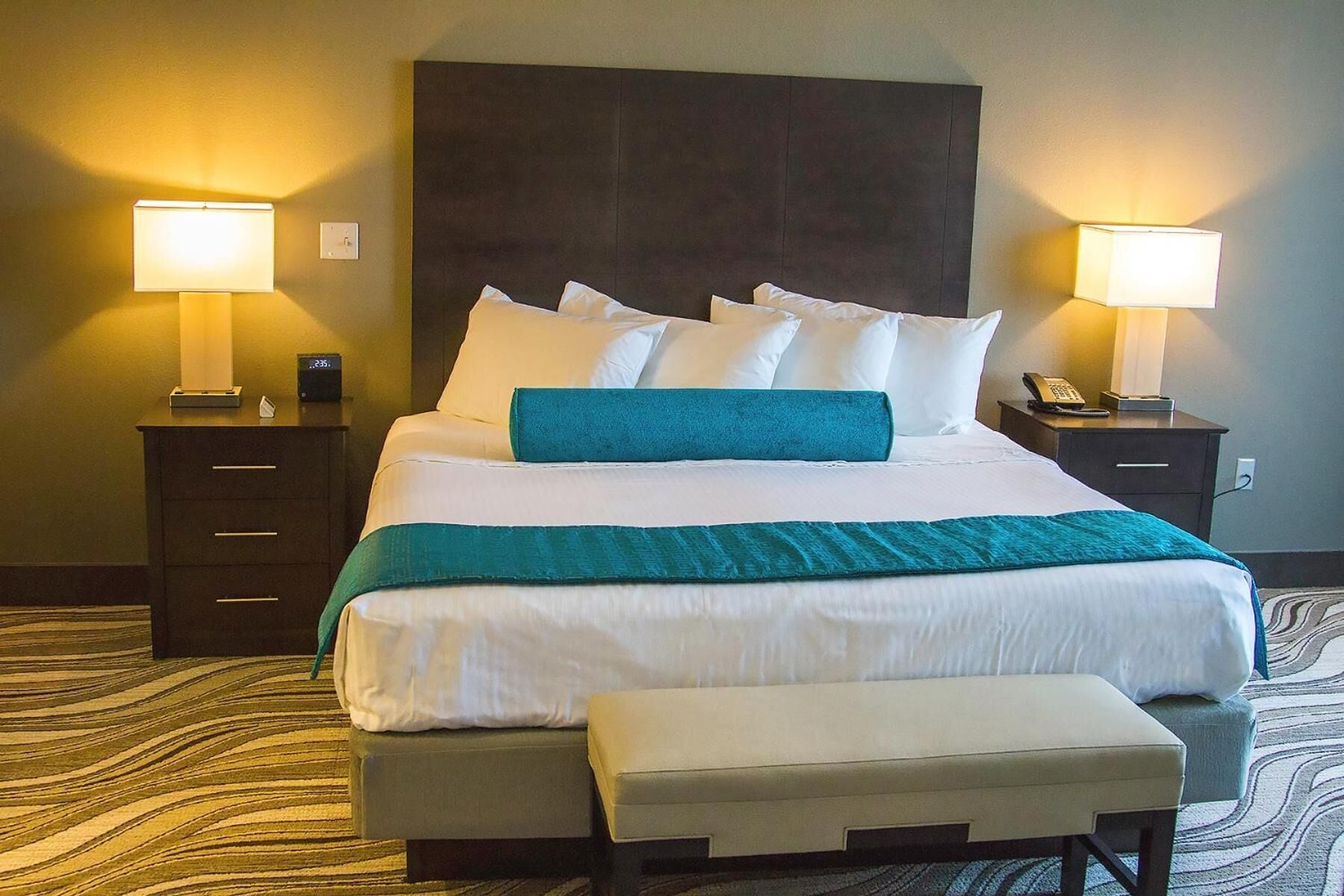 A hotel room with a large bed, two bedside tables, a nightstand, a bench, and two lamps.