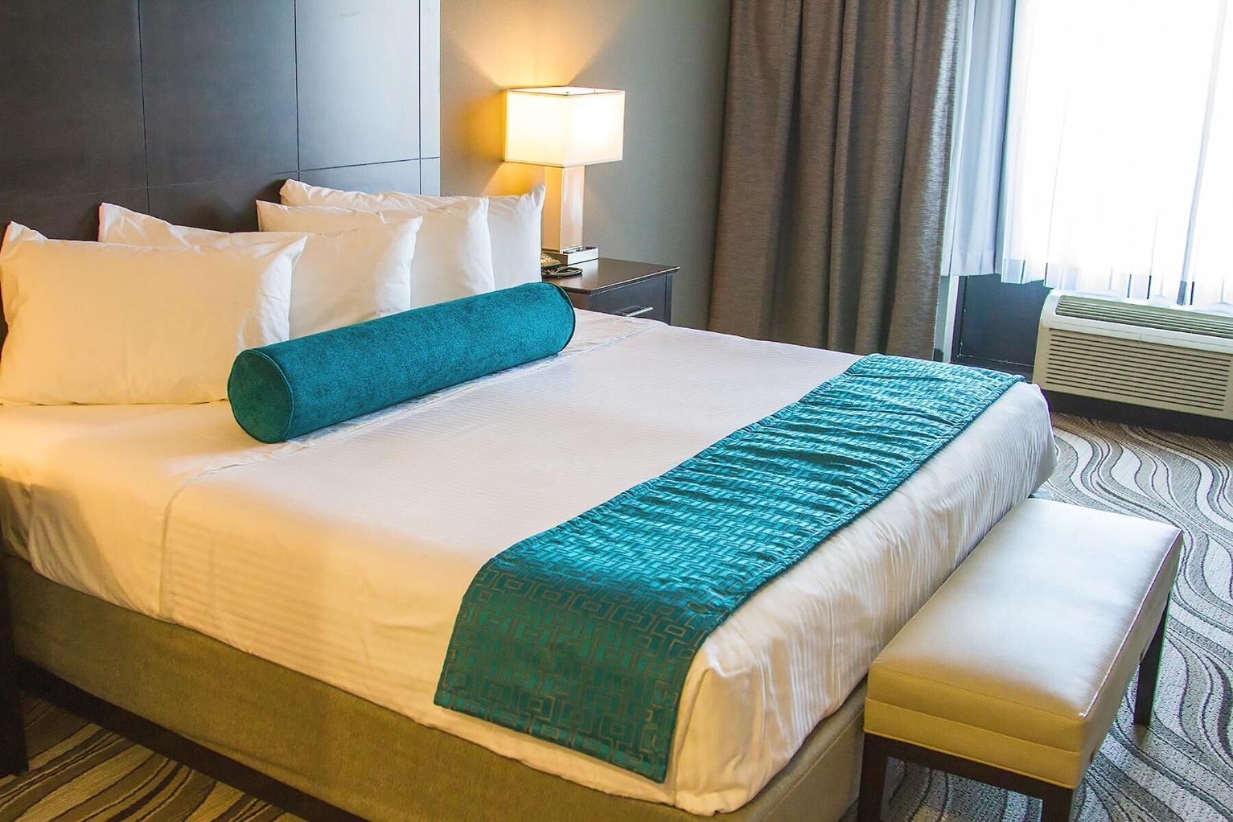 Habitación de hotel con una cama grande, una mesita de noche con lámpara, un banco y un equipo de aire acondicionado.