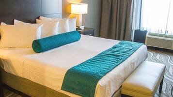 A hotel room with a large bed, a bedside table with a lamp, a bench, and an air conditioning unit.