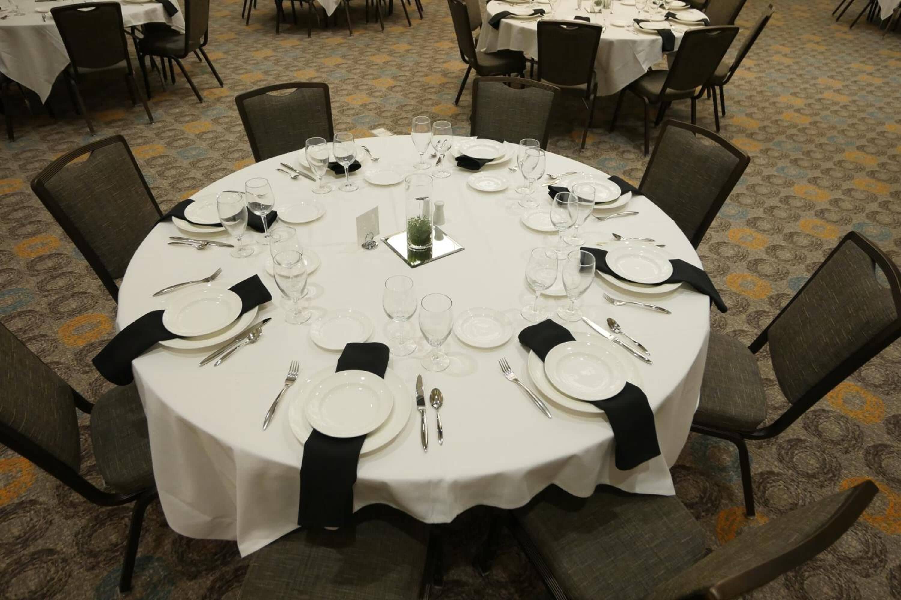 A round dining table set for eight with white tablecloths, black napkins, and silverware.