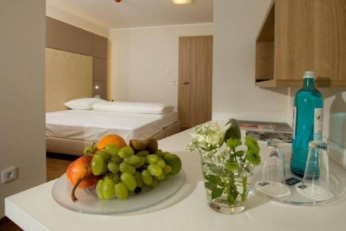 Une chambre d’hôtel avec un lit, un comptoir blanc sur lequel se trouve un panier de fruits, un vase avec des fleurs et une bouteille.