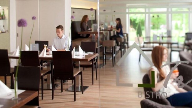 A modern bistro with wooden tables and chairs, a glass partition, and a view of greenery outside. - Hotel & Restaurant Bakenhof (Münster)
