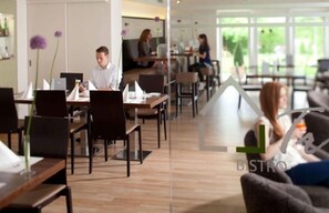 Un bistro moderne avec des tables et des chaises en bois, une cloison en verre et une vue sur la verdure à l’extérieur.
