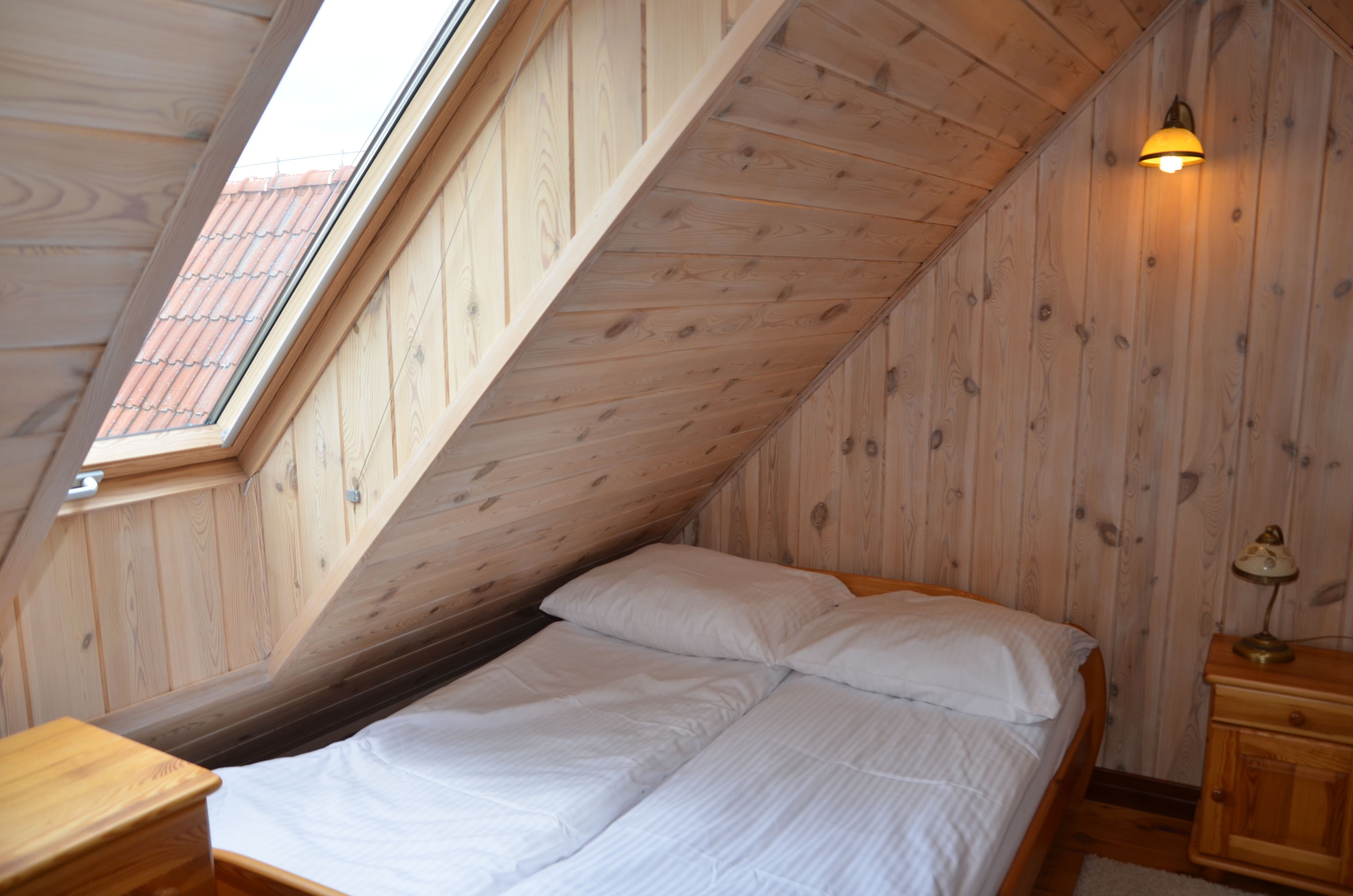 A room with a sloped ceiling, a bed with white bedding, a nightstand with a lamp, and a window with a view of a red-tiled roof.