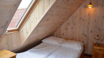 A room with a sloped ceiling, a bed with white bedding, a nightstand with a lamp, and a window with a view of a red-tiled roof.
