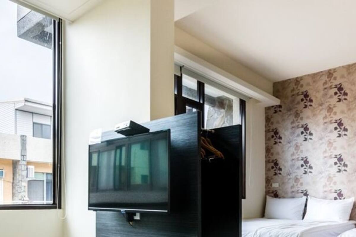 A hotel room with a bed, a TV, a wardrobe, and a window with a view of buildings.
