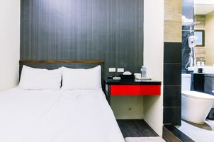 Una habitación de hotel con cama, mesita de noche, cajón rojo, espejo, lavabo e inodoro.
