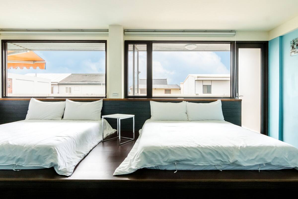 A hotel room with two single beds, a small table, and large windows offering a view of the cityscape.