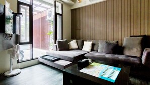 A modern living room with a sectional sofa, a glass coffee table with a magazine, a standing fan, and a large window with blinds.