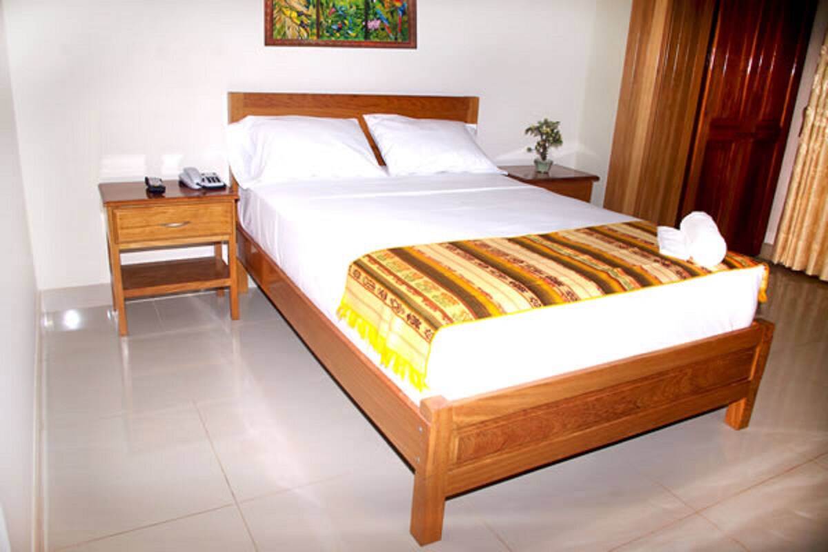 A hotel room with a wooden bed, white linens, a striped blanket, a bedside table with a phone and remote, and a painting on the wall.