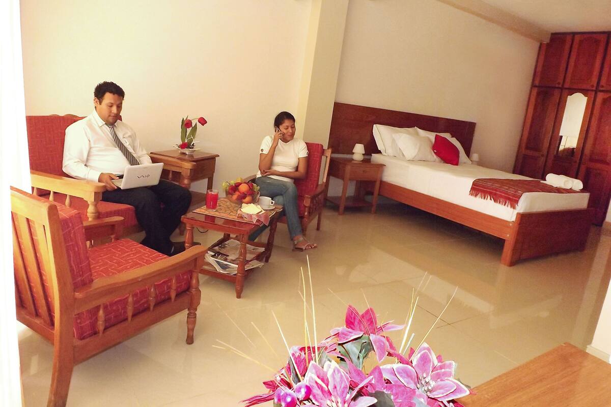 A hotel room with a bed, a sofa, a wooden coffee table, and a small table with a fruit bowl and a cup.