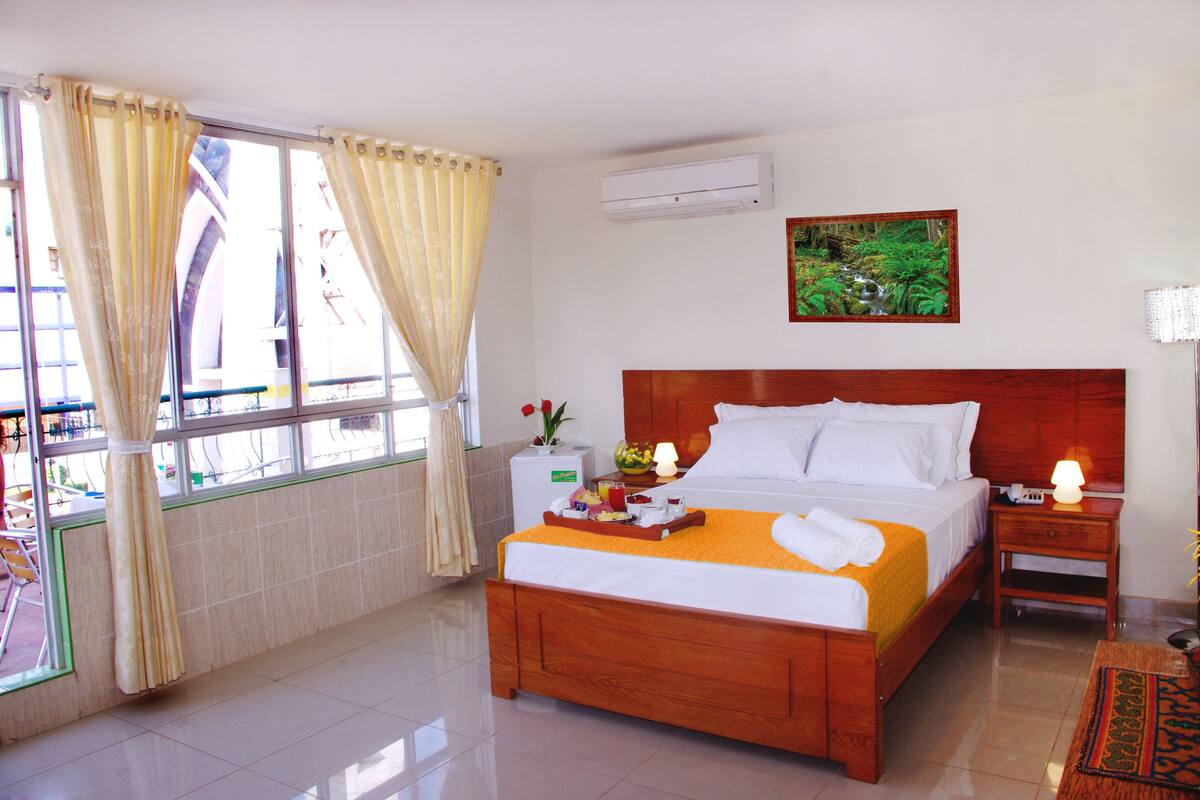 A hotel room with a wooden bed, a nightstand with a lamp, a window with curtains, a wall-mounted air conditioner, and a painting.