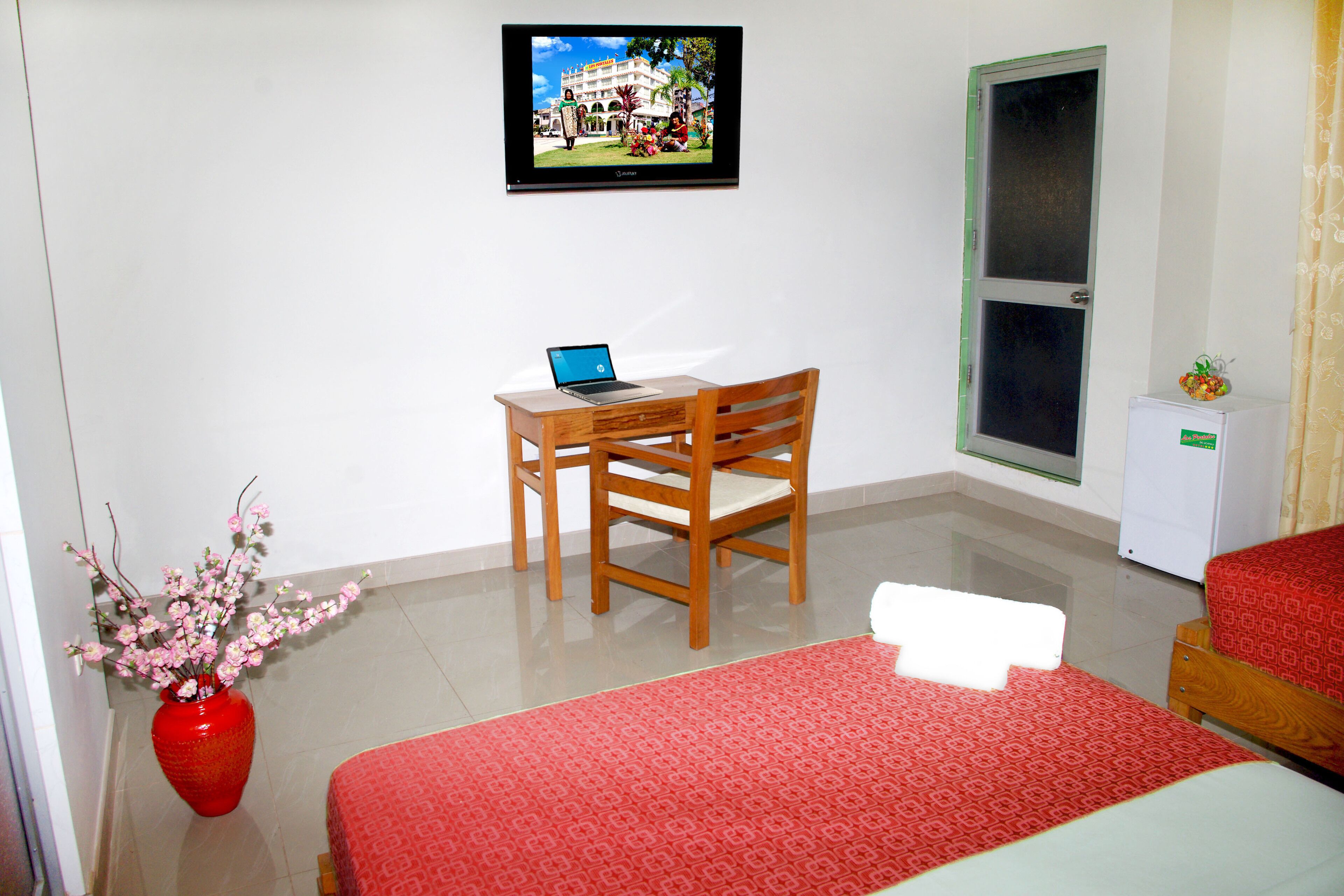 A hotel room with a bed, a desk with a laptop, a TV on the wall, a red vase with flowers, and a small refrigerator.