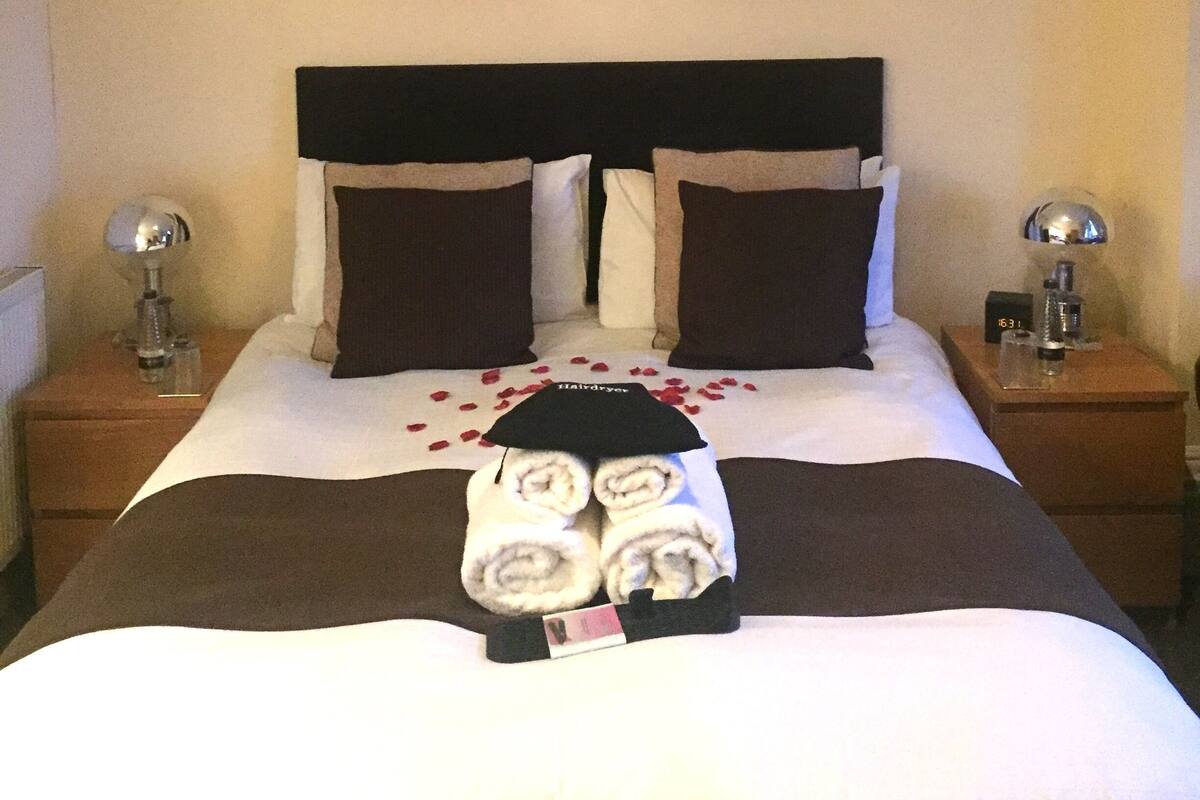 Un lit bien fait, décoré de pétales de rose, deux tableaux représentant des roses accrochés au mur et une horloge posée sur la table de chevet.