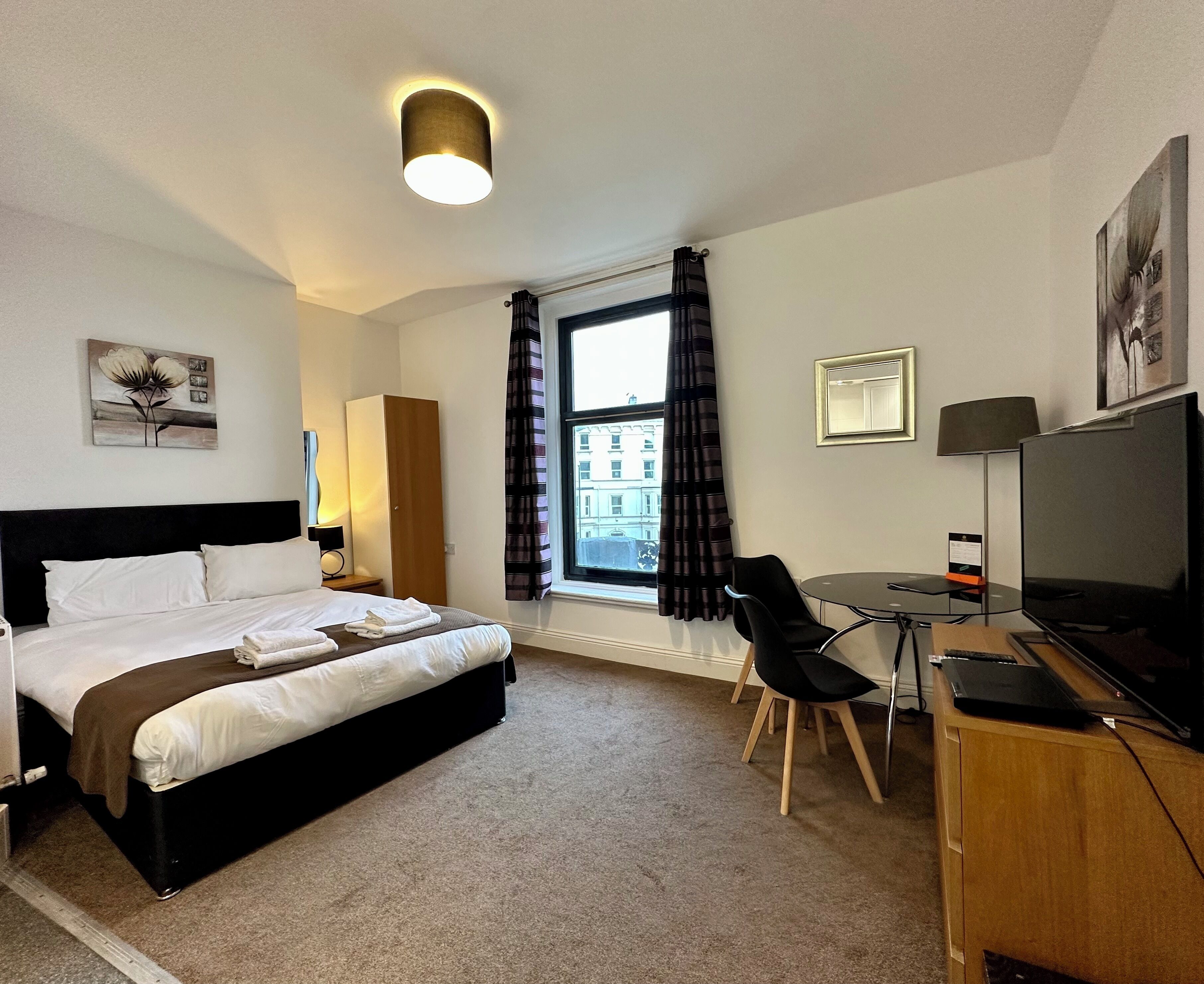 A hotel room with a bed, a desk with a chair, a TV, a window with curtains, and a painting on the wall.