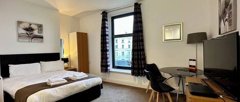 A hotel room with a bed, a desk with a chair, a TV, a window with curtains, and a painting on the wall.
