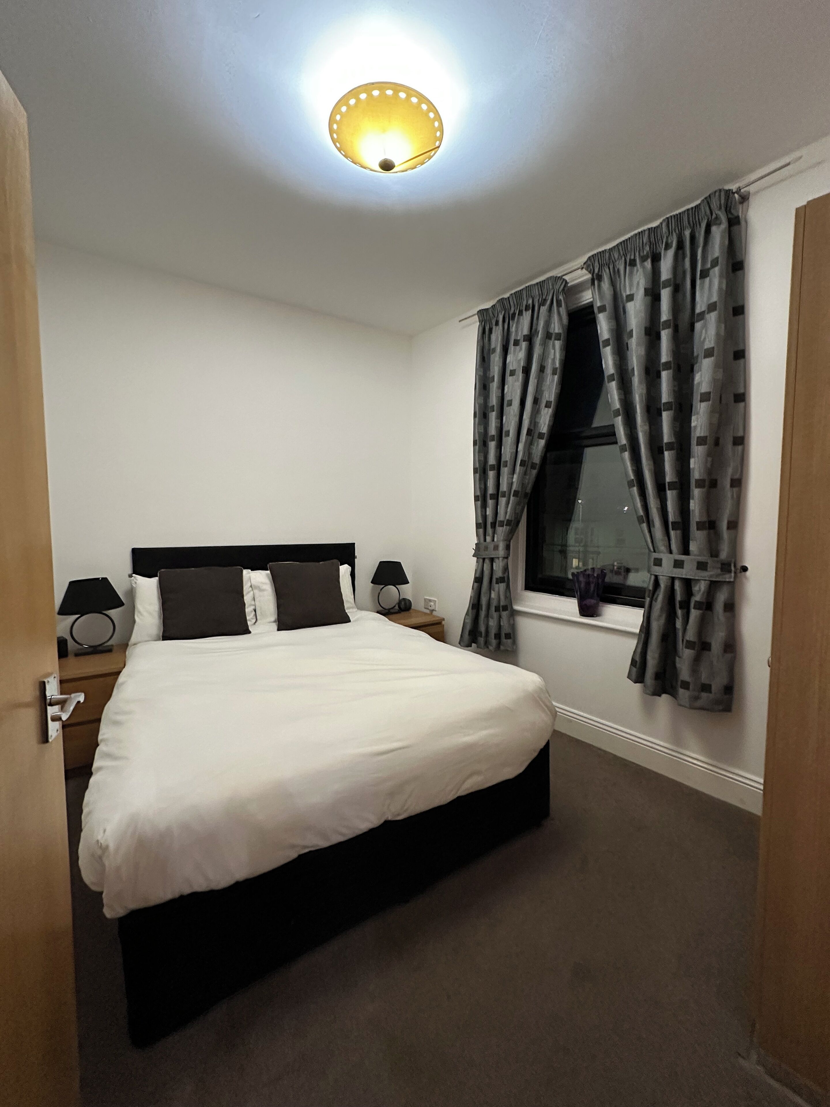 A neatly made bed in a hotel room with a window, curtains, and a ceiling light.
