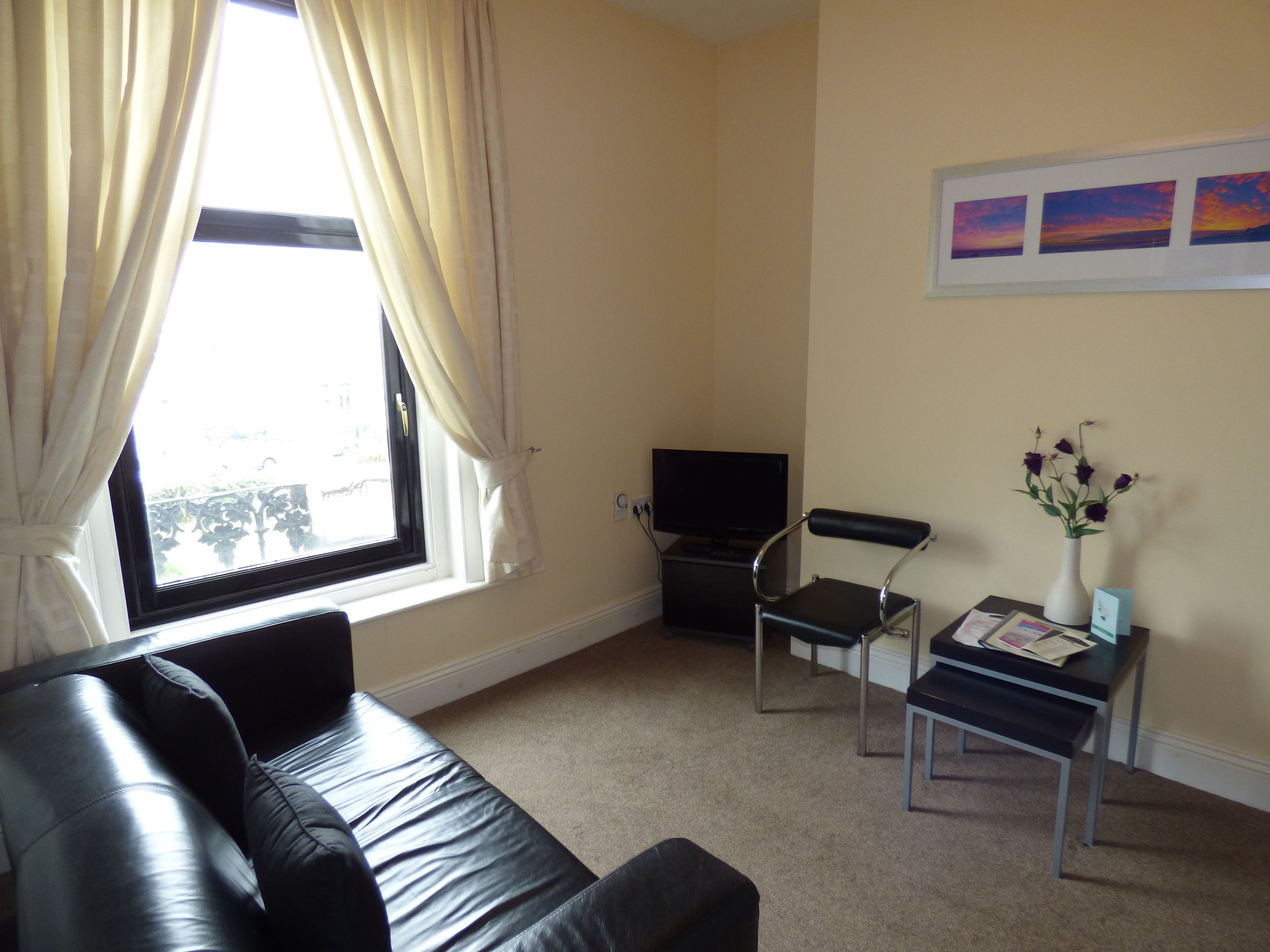 A living room with a black leather sofa, a small black table with a vase of flowers, a black chair, and a television on a stand.