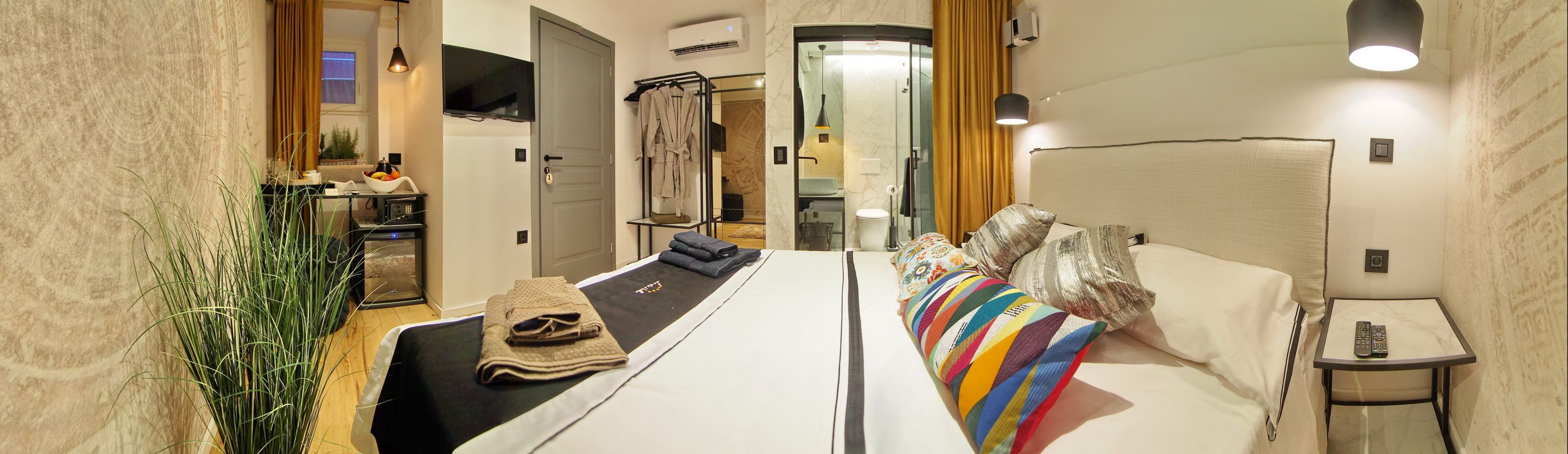 Une chambre d’hôtel avec un grand lit, une table de chevet avec une télécommande, un téléviseur fixé au mur et une salle de bain visible à travers une porte ouverte.