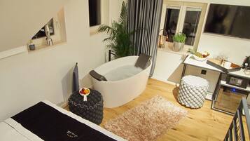 Un dormitorio moderno con una cama grande, una bañera, una pequeña kitchenette y una ventana con cortinas.