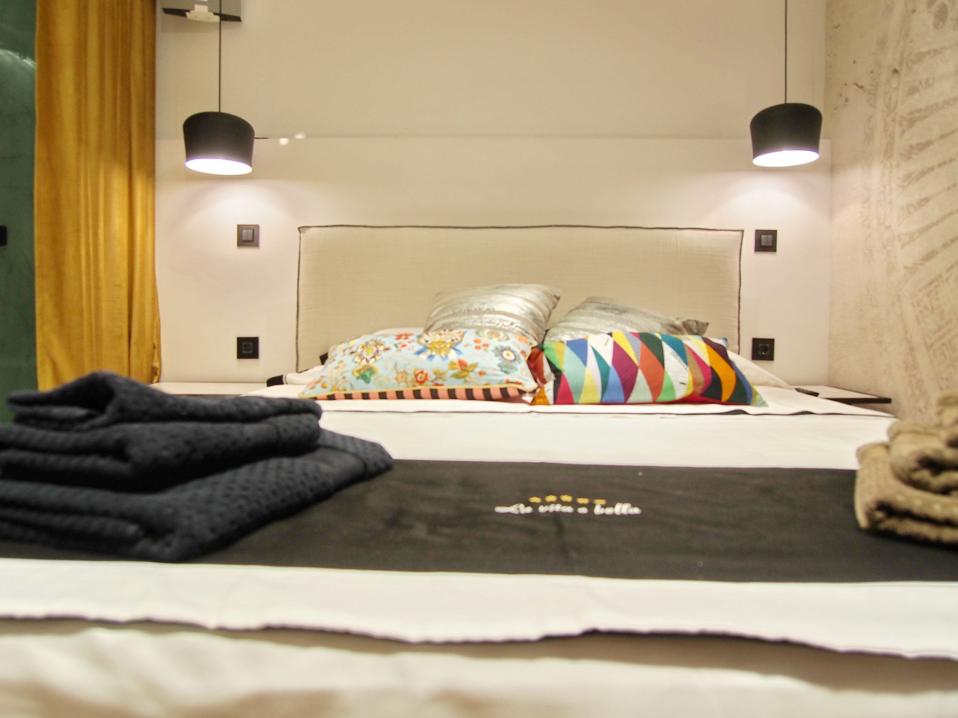 Ein ordentlich bezogenes Bett mit weißem Kopfteil, zwei Kissen mit floralen und bunten Mustern sowie einer schwarzen Decke mit kleinem Textlogo.