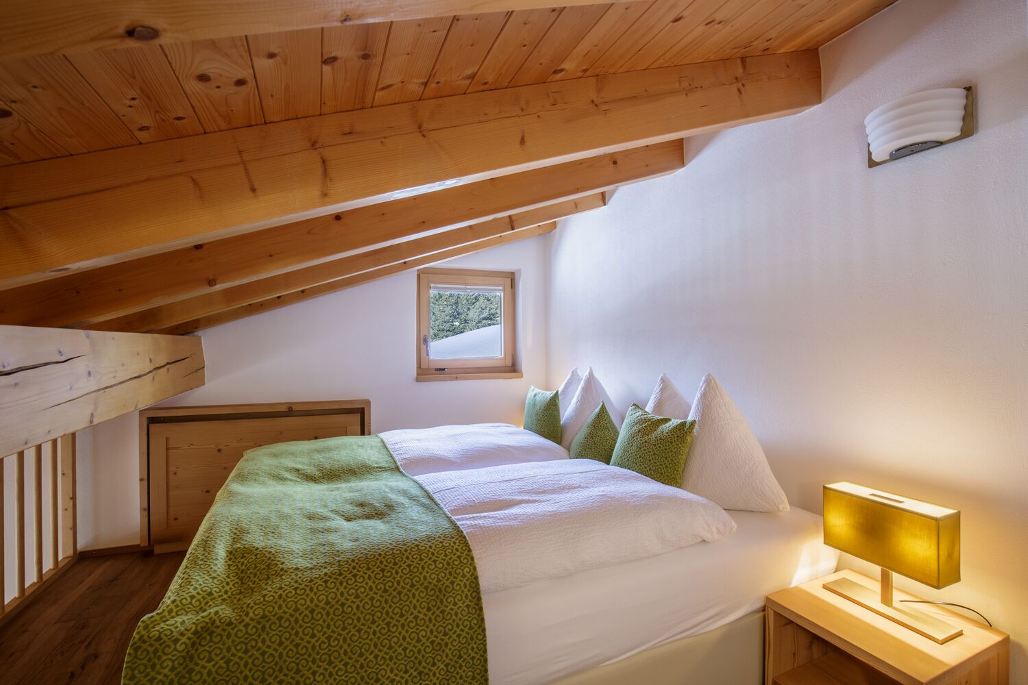 Ein gemütliches Schlafzimmer im Dachgeschoss, mit schrägen Wänden, einem Bett mit grüner Decke und weißen Kissen sowie einer Nachttischlampe.