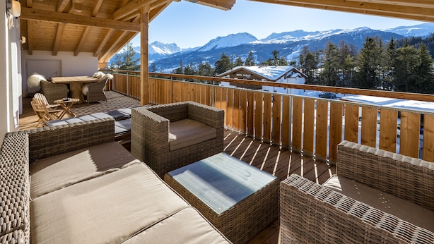 A wooden deck with wicker furniture, a table, and chairs, overlooking a snowy mountain range. - Hotel Sarain - Val Saraina Resort Lenzerheide (Lantsch-Lenz)